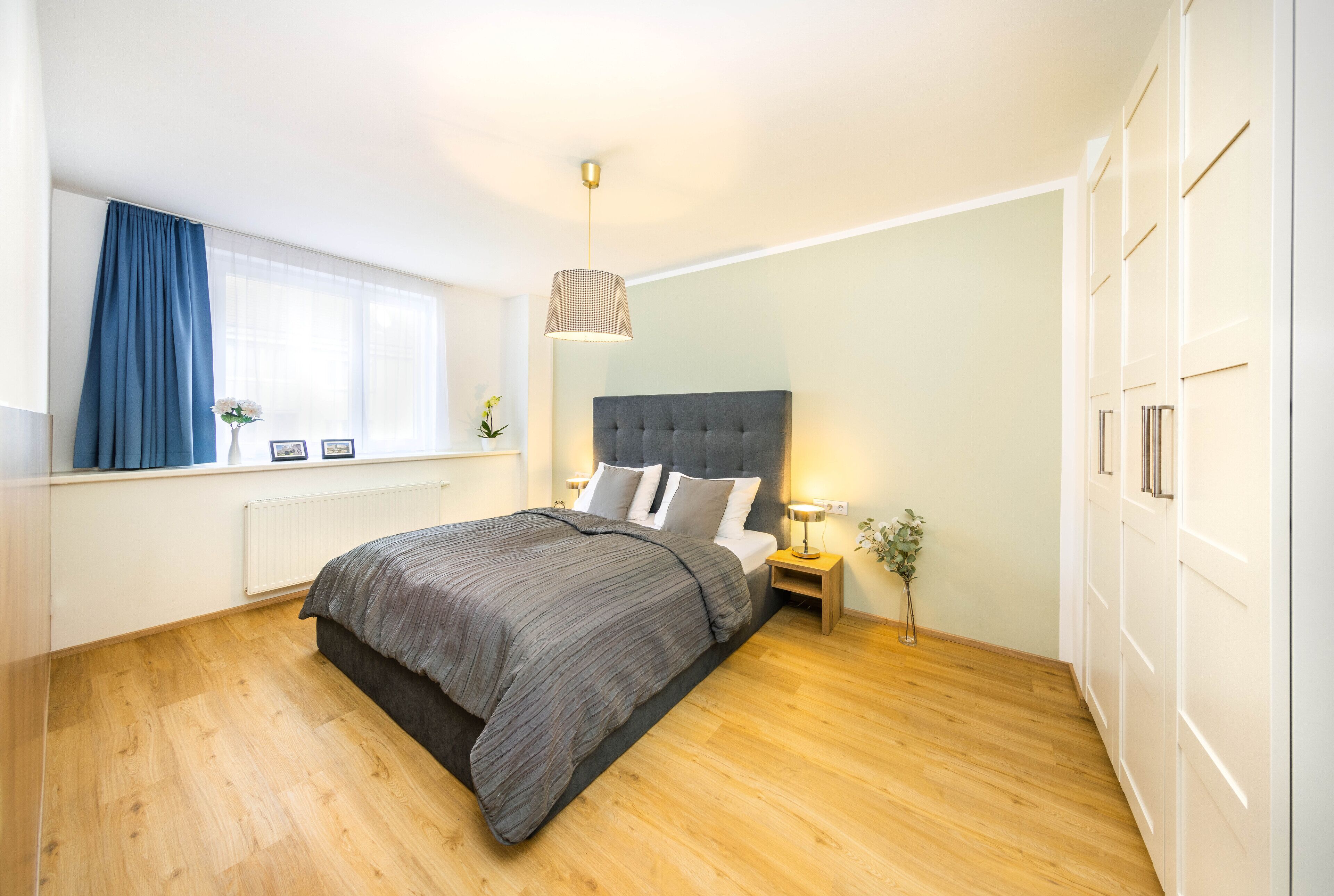 Een slaapkamer met een groot bed, een nachtkastje met lamp, een raam met gordijnen en een kledingkast.