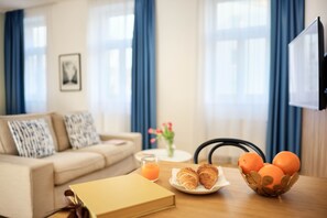 A living room with a sofa, a coffee table, a TV, a bowl of oranges, a plate with croissants, and a book.