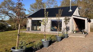 A modern wooden house with large windows, a covered porch, and a gravel driveway.