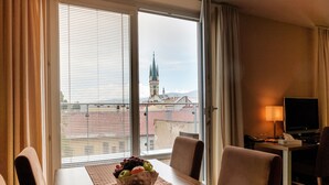 Habitación de hotel con mesa de comedor, sillas, frutero, televisor y vista a un paisaje urbano con una torre de iglesia.