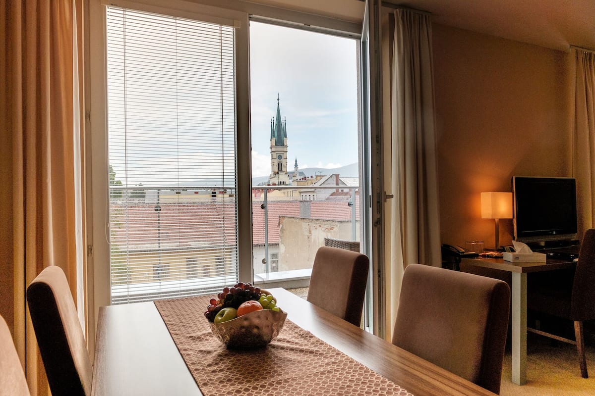 A hotel room with a dining table, chairs, a fruit bowl, a TV, and a view of a cityscape with a church tower.