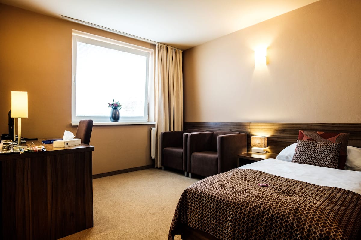 A hotel room with a bed, desk, chairs, a window with curtains, a nightstand, and a lamp.