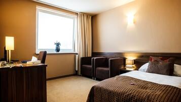 A hotel room with a bed, desk, chairs, a window with curtains, a nightstand, and a lamp.
