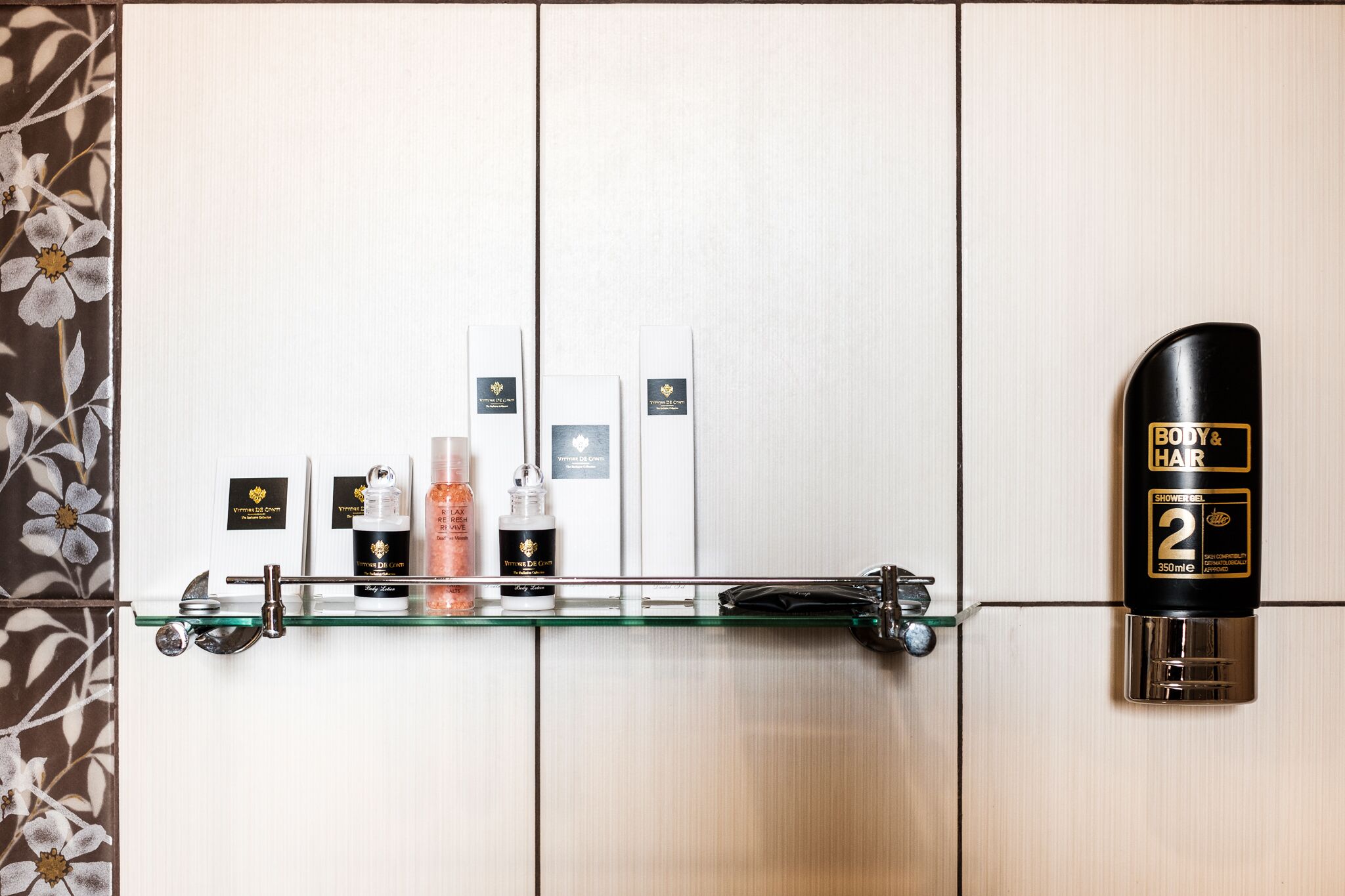 Ein Badezimmer mit einer Glasablage, auf der Pflegeprodukte stehen, einer schwarzen Flasche mit BODY & HAIR Duschgel und einer Wand mit Blumenmuster.