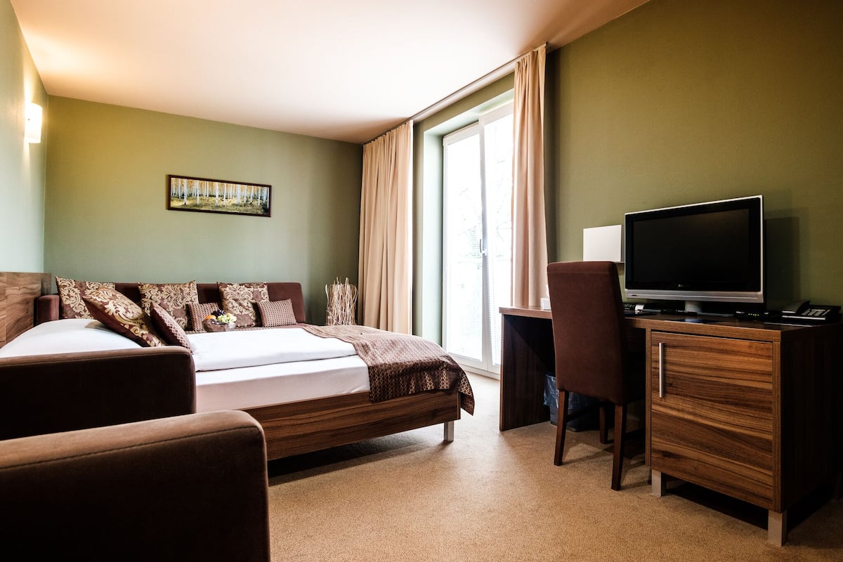 A hotel room with a bed, a desk with a TV, a chair, and a window with curtains.