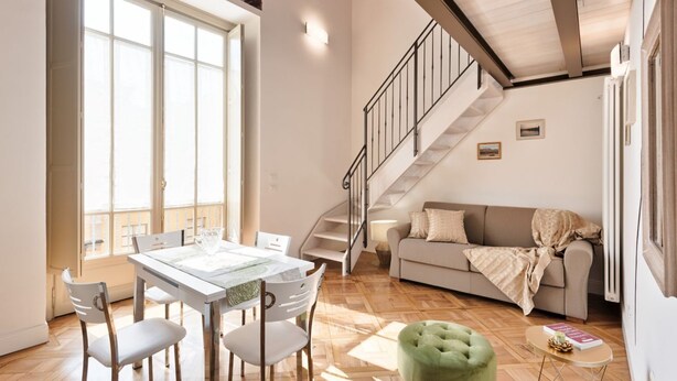 Een ruime woonkamer met een eettafel, stoelen, een bank, een groene poef en een trap.