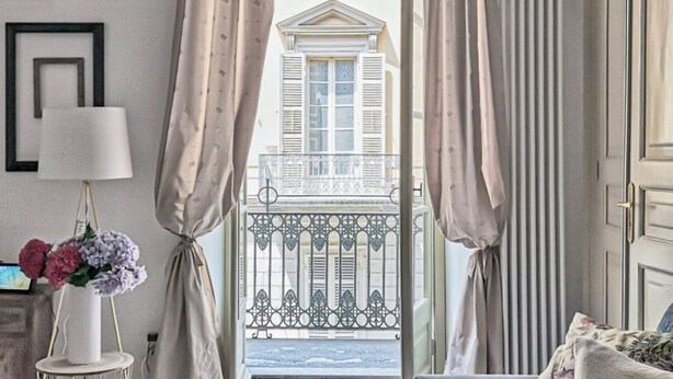 Une pièce avec vue sur un balcon, un vase blanc rempli de fleurs et une lampe sur une table d’appoint.