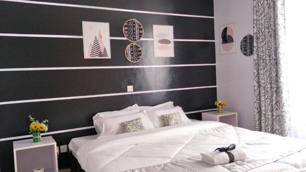 A modern bedroom with a black and white striped wall, a neatly made bed with white linens, and decorative wall art. - Cozy Room JKIA (Mlolongo)
