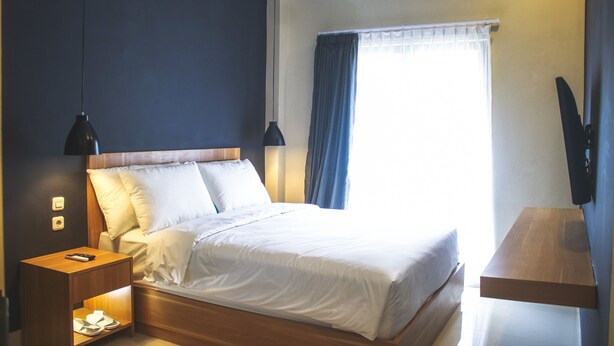 A neatly made bed with white linens, a wooden headboard, a bedside table with a lamp, and a window with blue curtains.
