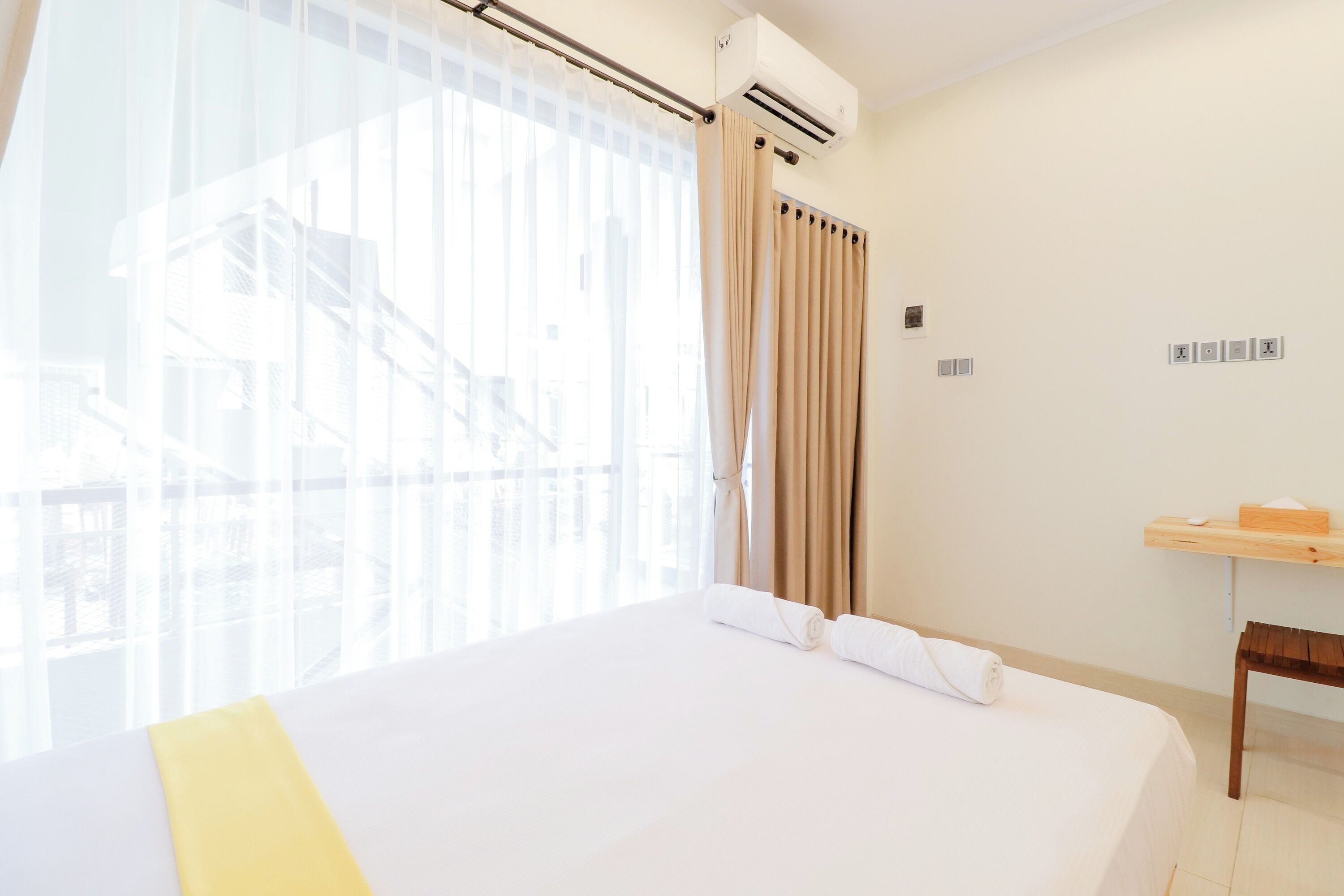 A hotel room with a large bed, a wooden nightstand, a window with sheer curtains, and an air conditioning unit.