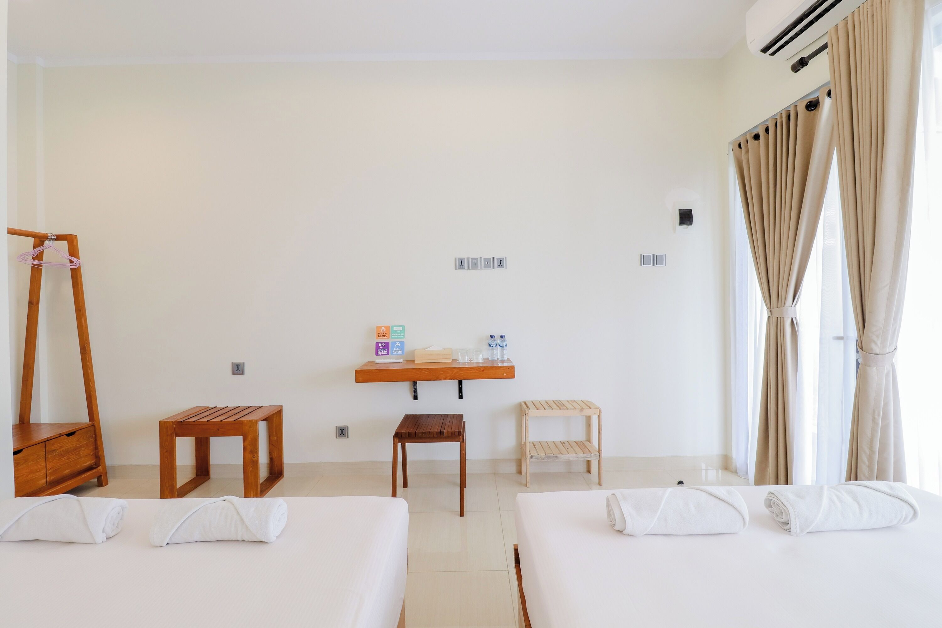 A hotel room with two beds, a wooden table, a small stool, a mirror, and a shelf with amenities.
