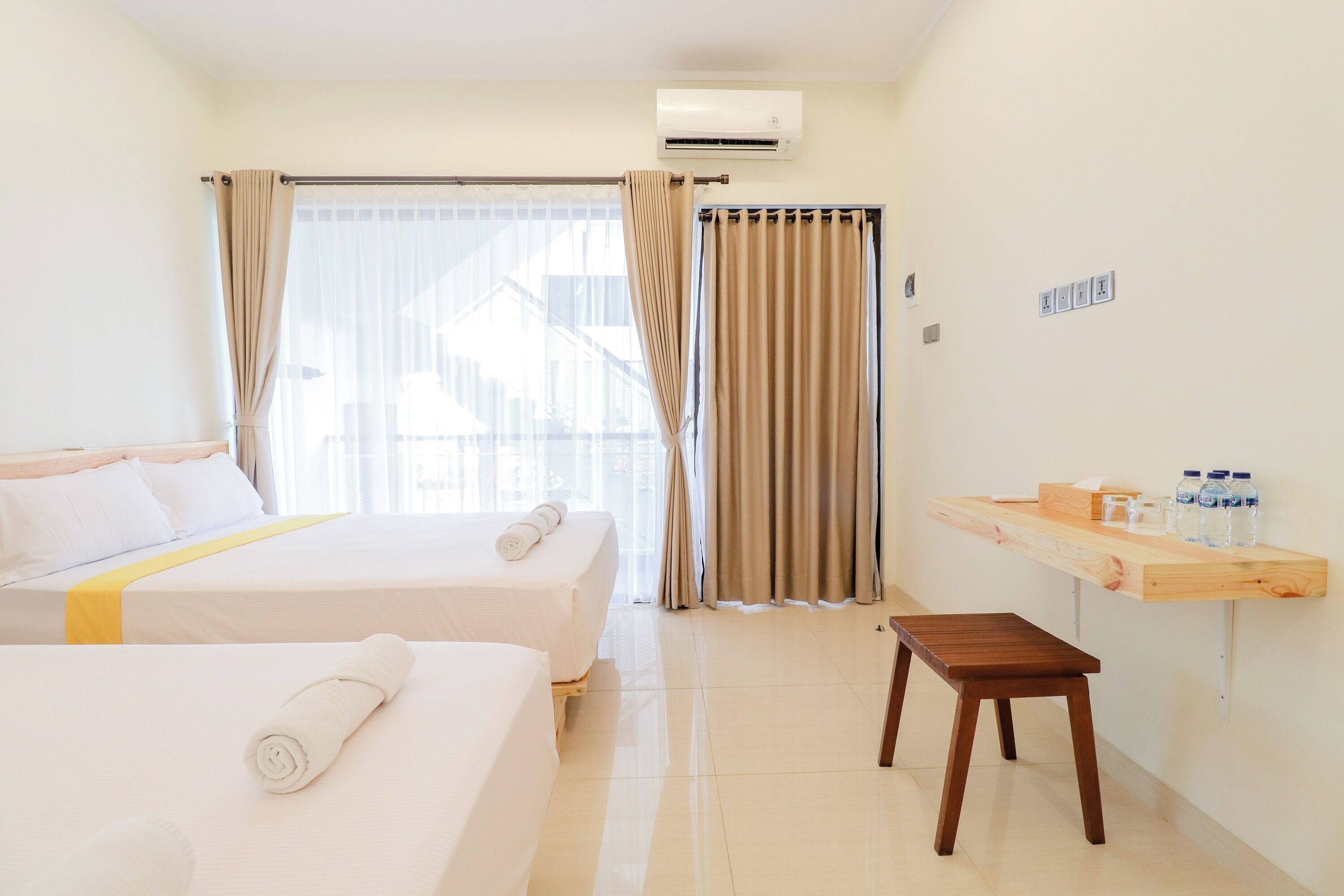 A hotel room with two beds, a wooden bench, a small table with toiletries, and a window with curtains.