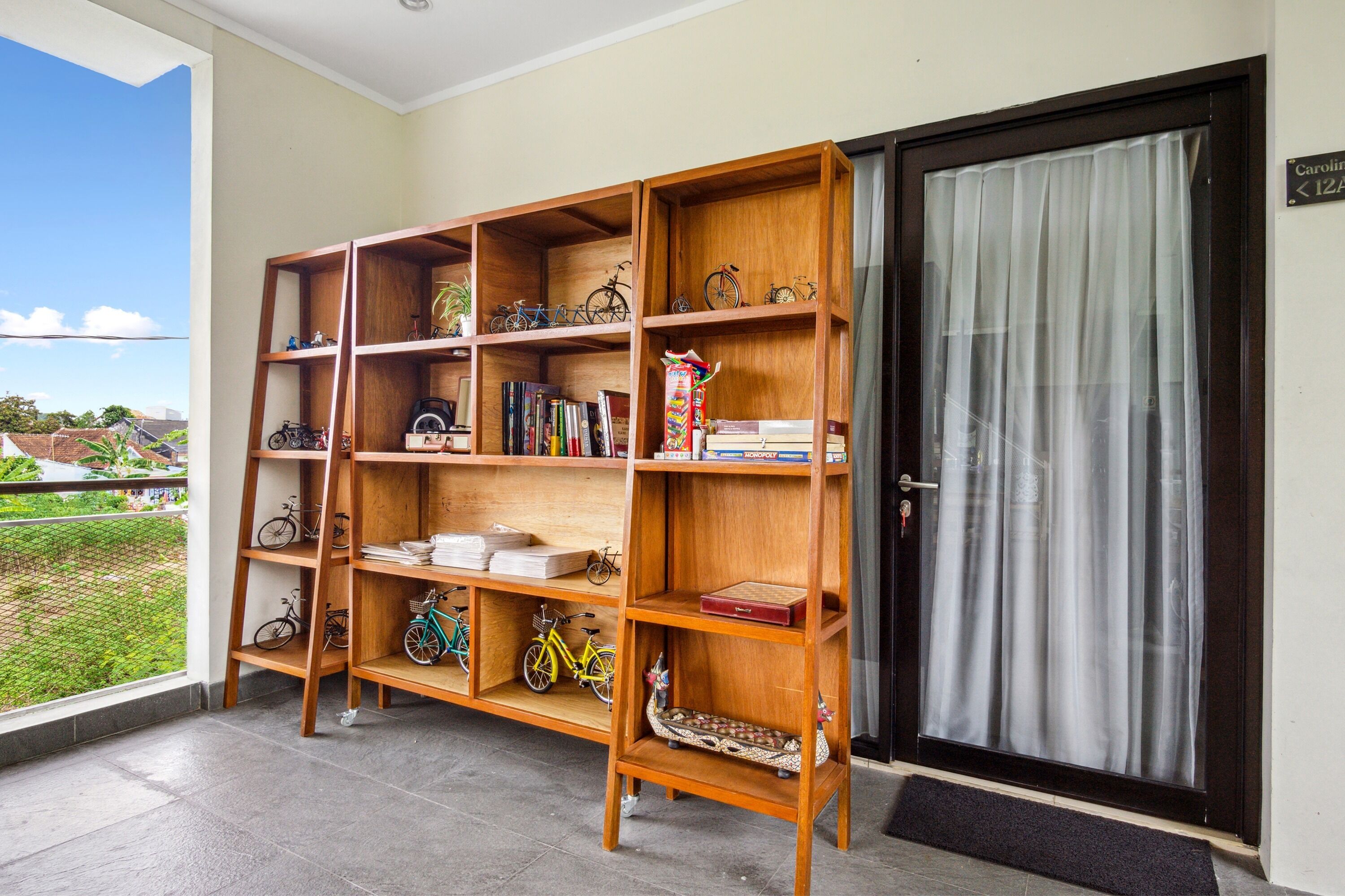 A wooden bookshelf with bicycles and books, a door with a sign, and a view of greenery outside.