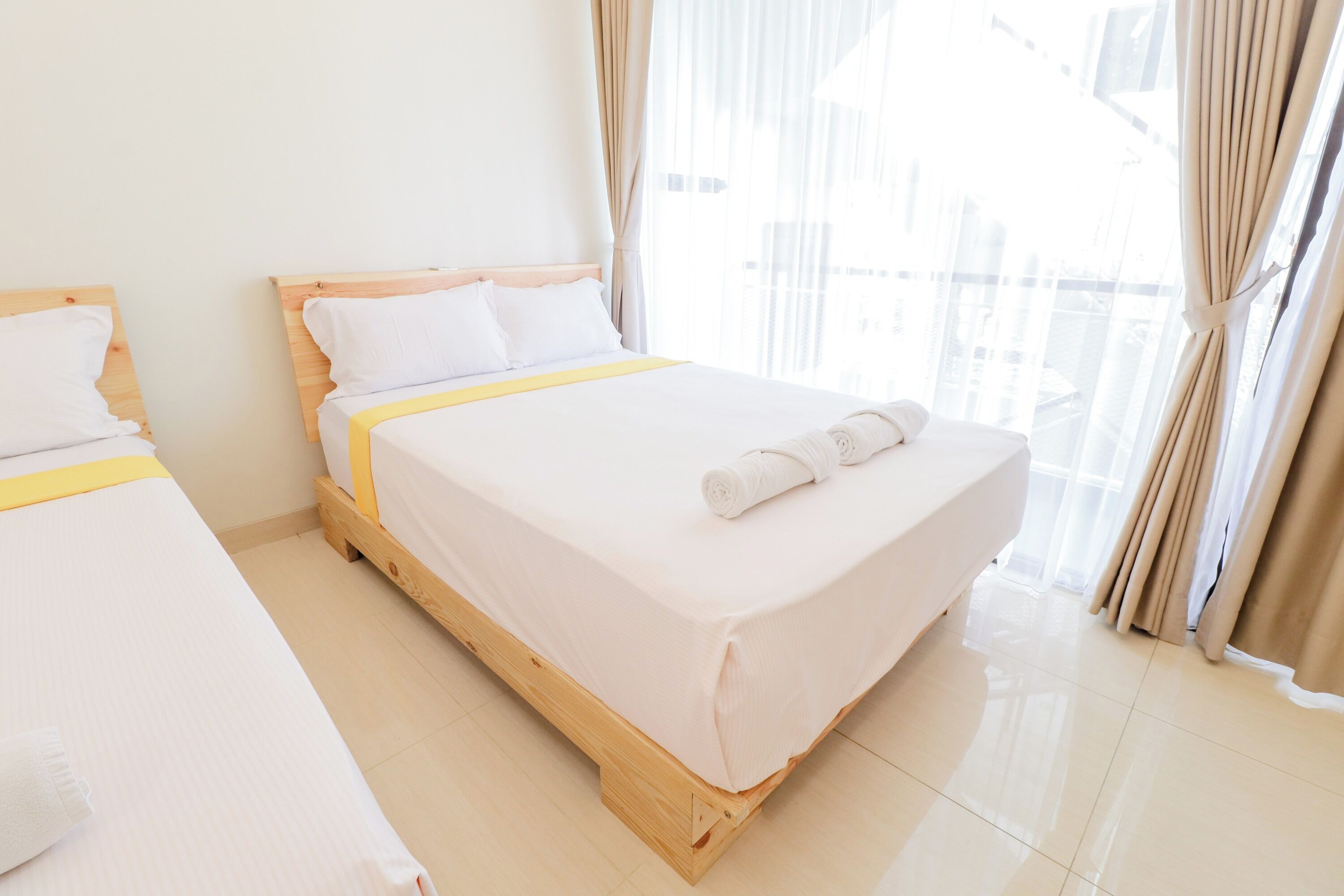 A hotel room with two beds, wooden headboards, white linens, and a view through sheer curtains.