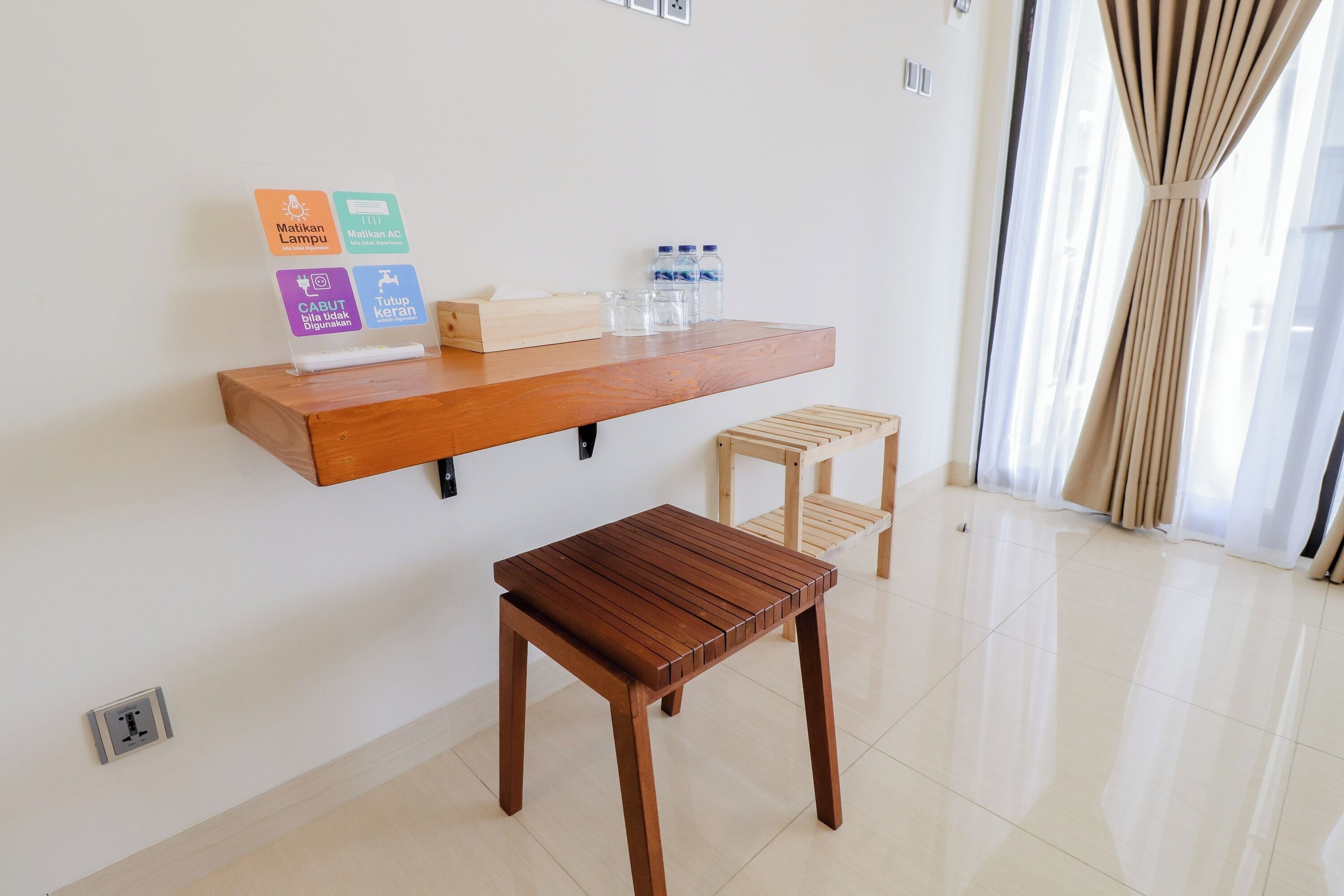 A clean, modern room with a wooden table and bench, a shelf with amenities, and a large window with curtains.