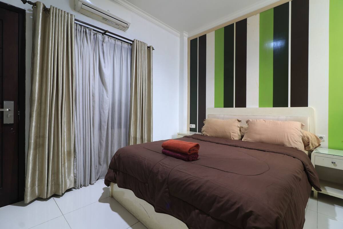 A bedroom with a bed, curtains, a nightstand, and a wall with vertical stripes.