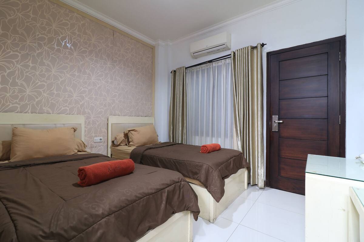 A hotel room with two beds, beige patterned wallpaper, an air conditioning unit, a dark wooden door, and a glass desk.