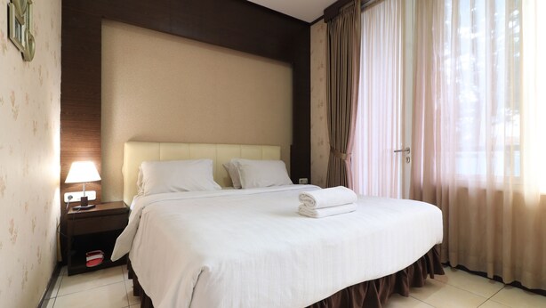 A neatly made bed with white linens, a bedside lamp, and a nightstand with a red object on it. - I Residence Bandung (Bandung)