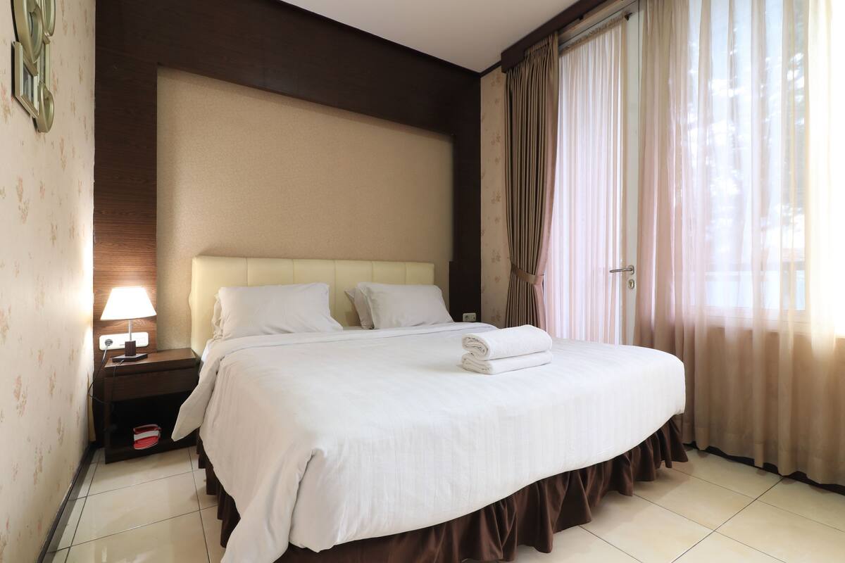 A neatly made bed with white linens, a bedside lamp, and a nightstand with a red object on it.
