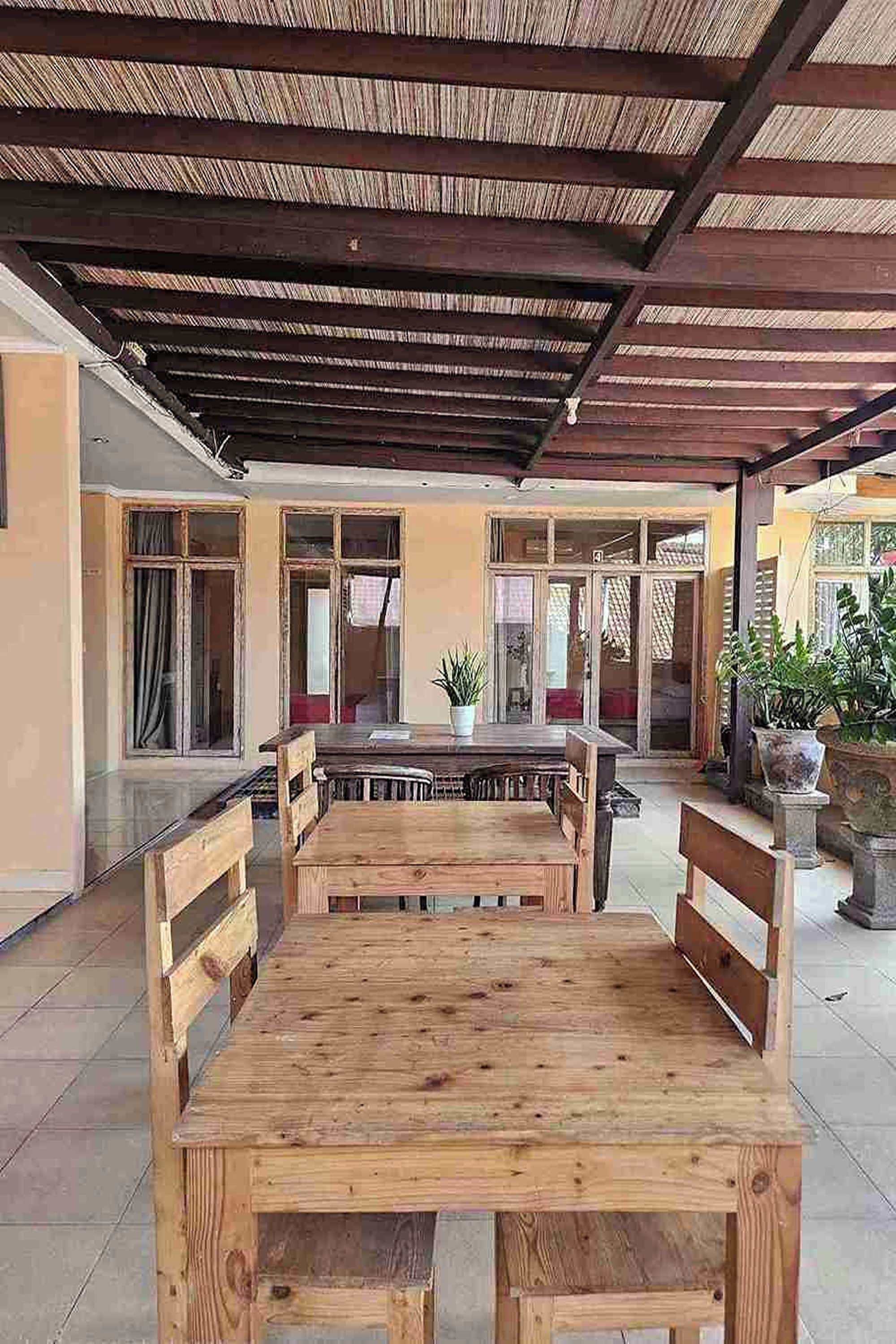 An outdoor patio with a wooden table and benches, surrounded by potted plants and a covered area with a sloped roof.