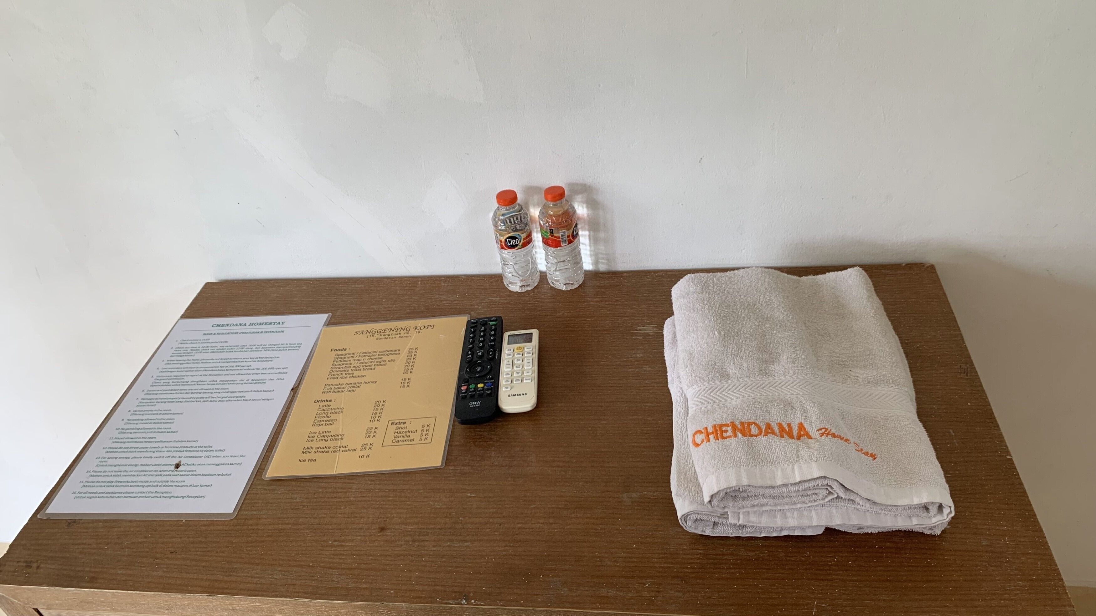 A wooden table with a menu, two water bottles, a remote control, a Samsung calculator, and a folded towel with "CHENDANA" embroidered on it.