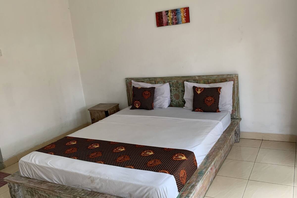 A single bed with a white mattress and a patterned bedspread, two pillows with a circular design, a small wooden nightstand, and a plain wall with a single piece of artwork.
