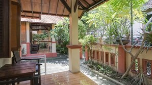 Un patio al aire libre con una mesa y sillas de madera, piso de baldosas y vista a un jardín con plantas diversas y un edificio al fondo.