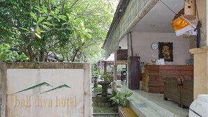Entrada de hotel con un cartel que dice "bali diva hotel", un camino empedrado, un pequeño jardín con una fuente y un mostrador de recepción con un reloj y una estatua.