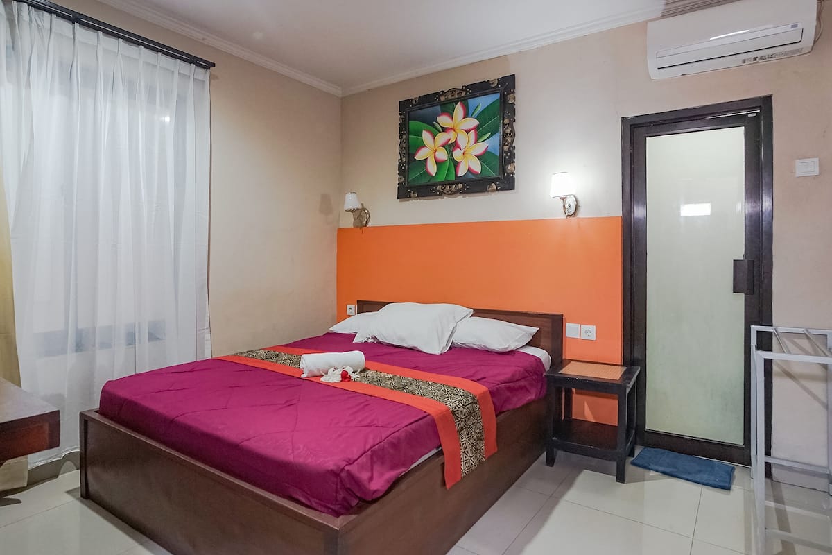 A hotel room with a bed, a nightstand, a desk, a chair, a window with curtains, an air conditioning unit, and a door.