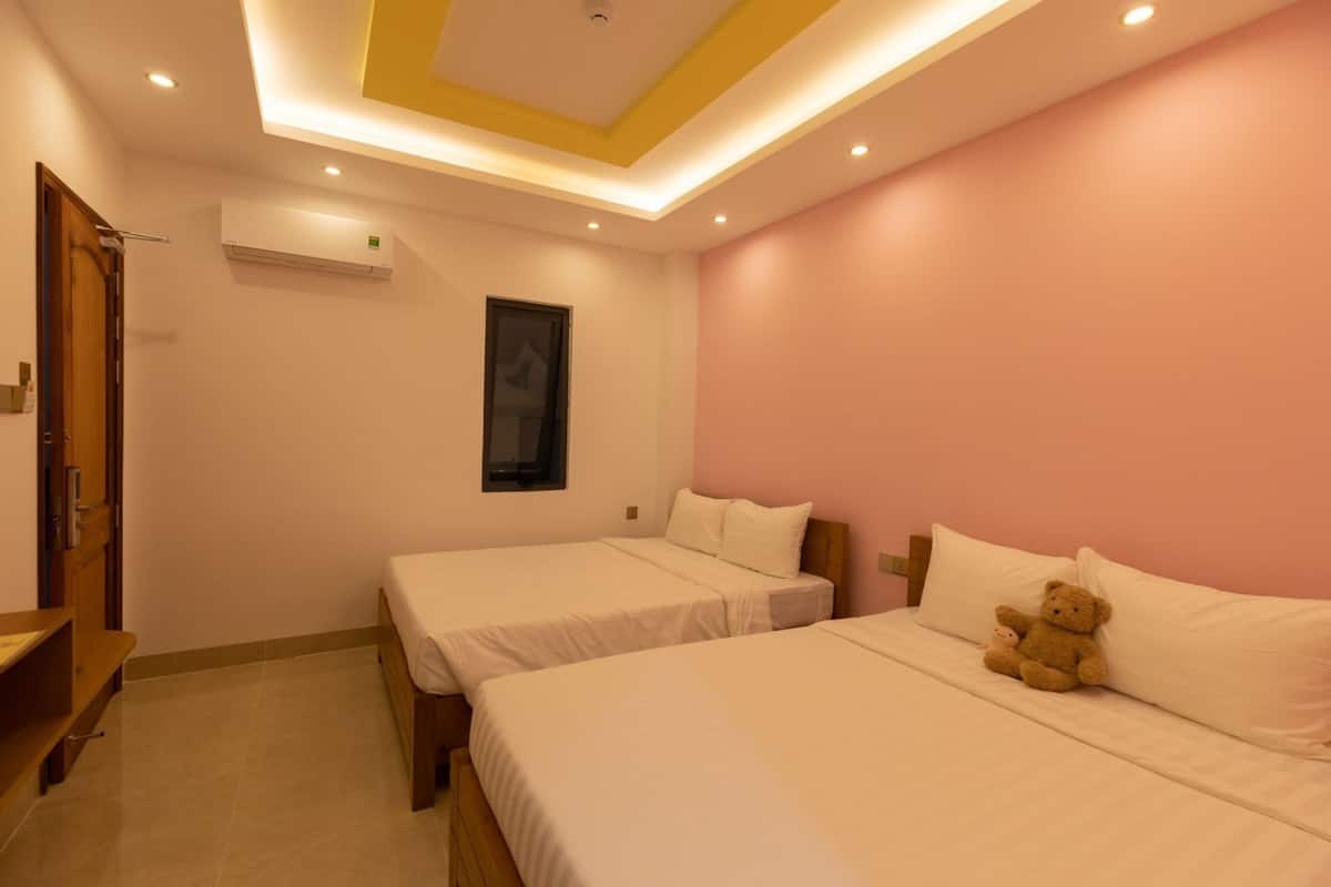 A hotel room with two beds, a wooden bench, a teddy bear, and a ceiling with recessed lighting.