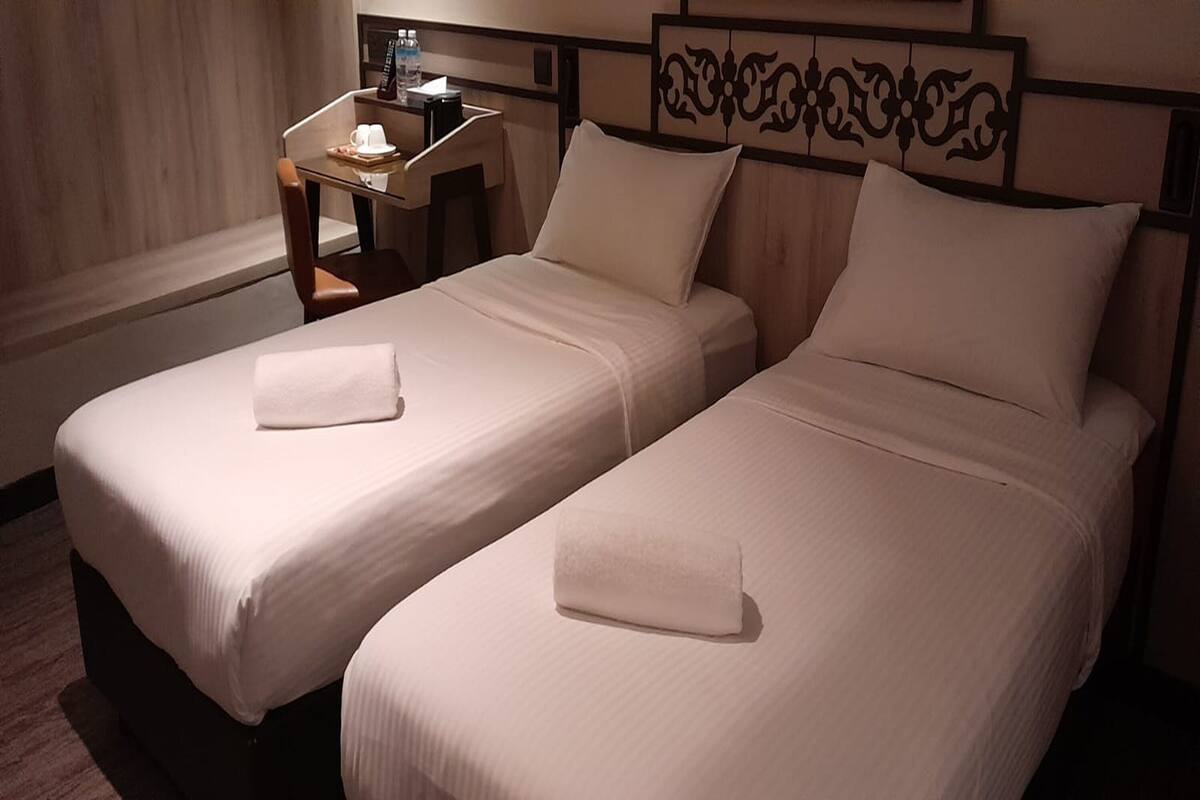 Deux lits simples avec du linge de lit et des oreillers blancs, une petite table avec une tasse et une bouteille, et une tête de lit décorative.