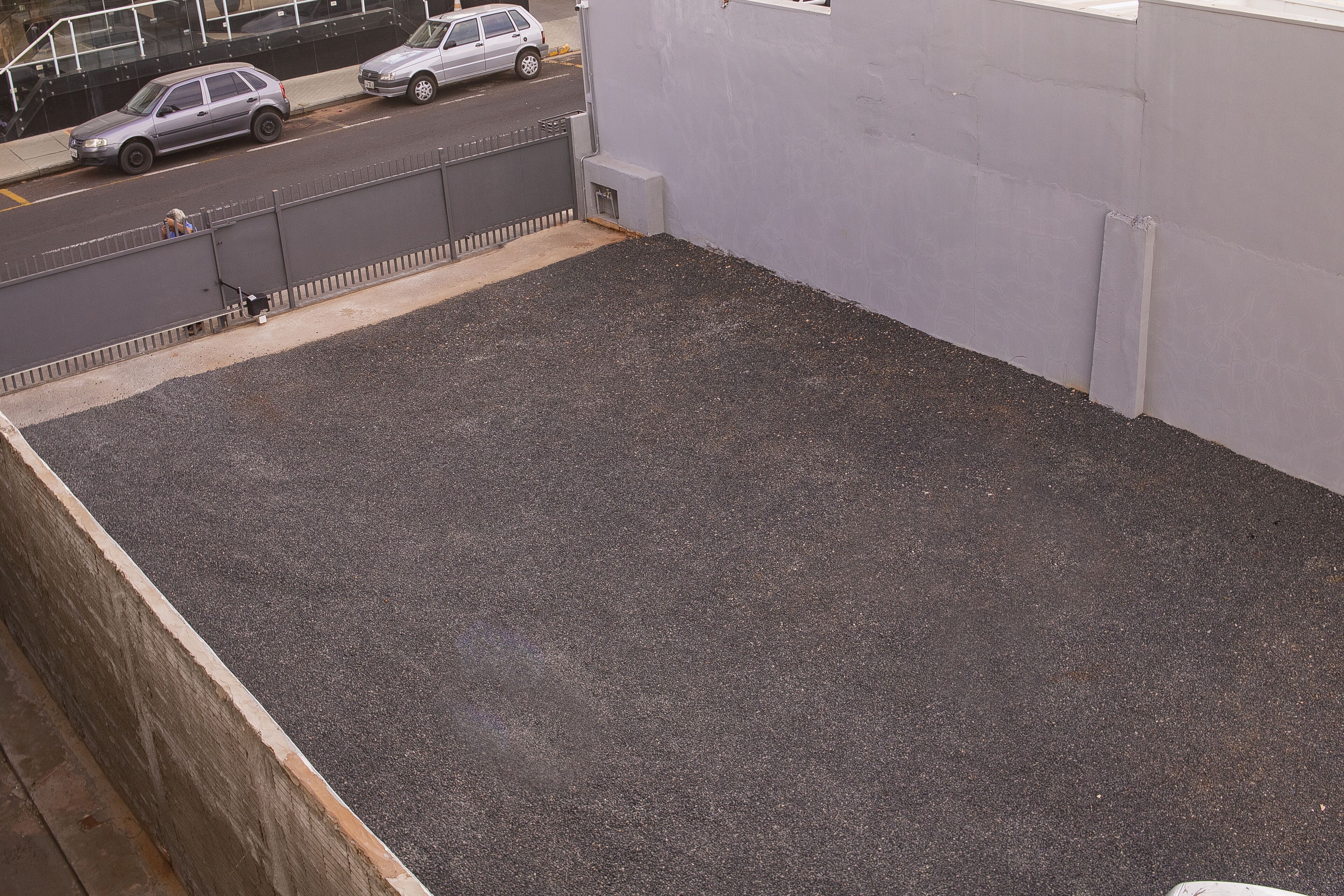 A rooftop with a gravel surface, a metal fence, and a building in the background.