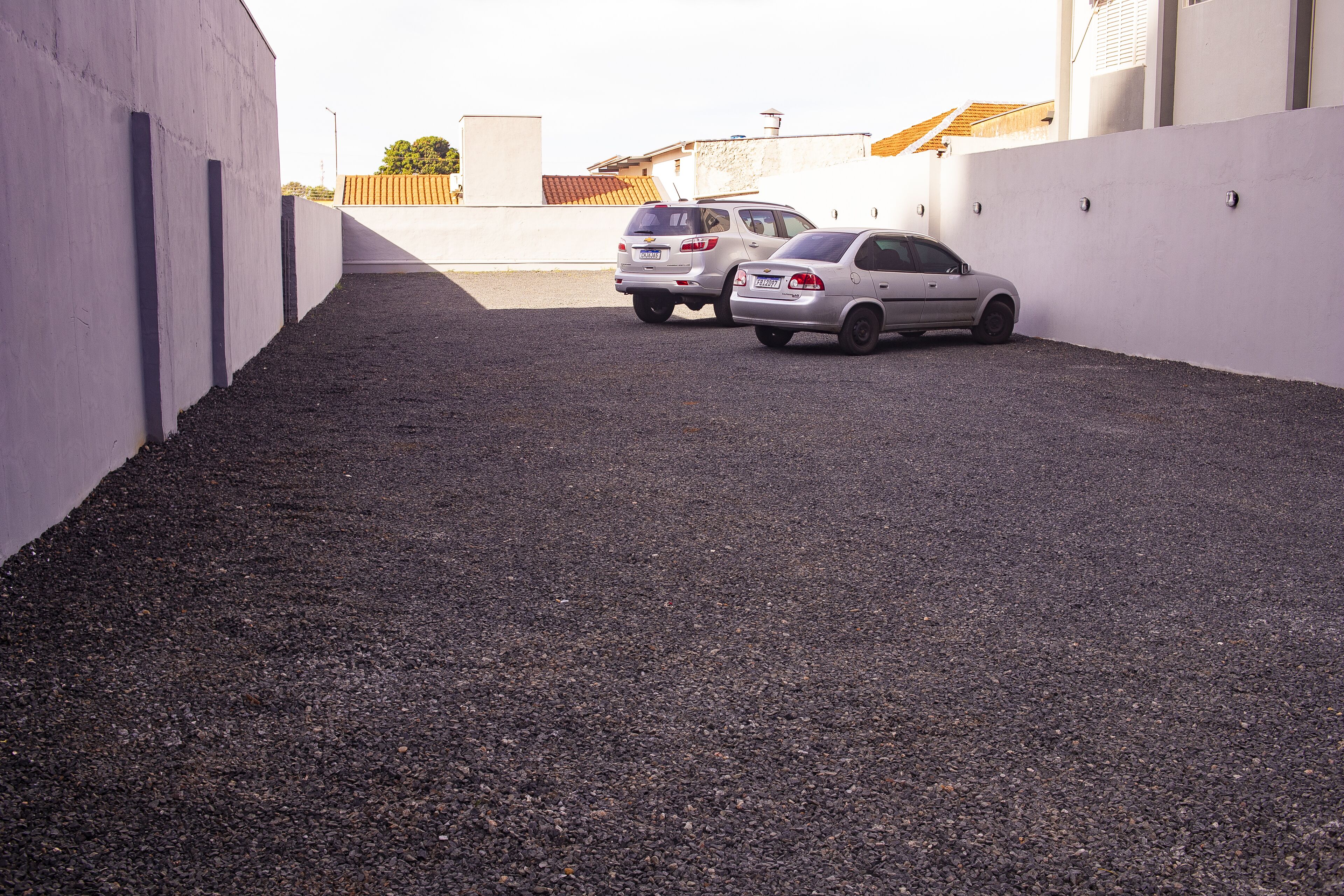 Un área de estacionamiento con dos coches aparcados uno tras otro, rodeada por un muro blanco y un suelo de grava.