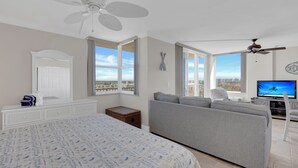 Ein geräumiges Schlafzimmer mit einem großen Bett, einem grauen Sofa, einem Fernseher und Blick auf die Stadt durch große Fenster.