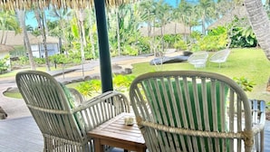 A covered outdoor seating area with wicker chairs and a table, overlooking a tropical garden with palm trees and a thatched-roof structure. - Motu Beachfront Art Villas (Rarotonga)