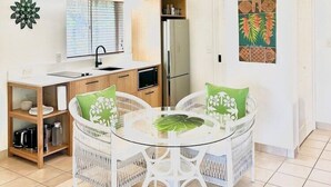 A compact kitchen with a refrigerator, microwave, and wooden cabinets. There is a dining area with a glass table and white wicker chairs, and a wall-mounted light fixture. - Motu Beachfront Art Villas (Rarotonga)