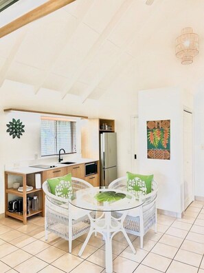 A compact kitchen with a refrigerator, microwave, and wooden cabinets. There is a dining area with a glass table and white wicker chairs, and a wall-mounted light fixture. - Motu Beachfront Art Villas (Rarotonga)
