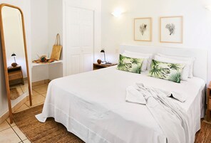 A neatly arranged bedroom with a large bed, a bedside table with a lamp, a mirror, and two framed botanical prints on the wall.