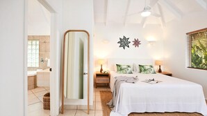 A bedroom with a bed, bedside tables, a mirror, and a window with blinds. - Motu Beachfront Art Villas (Rarotonga)