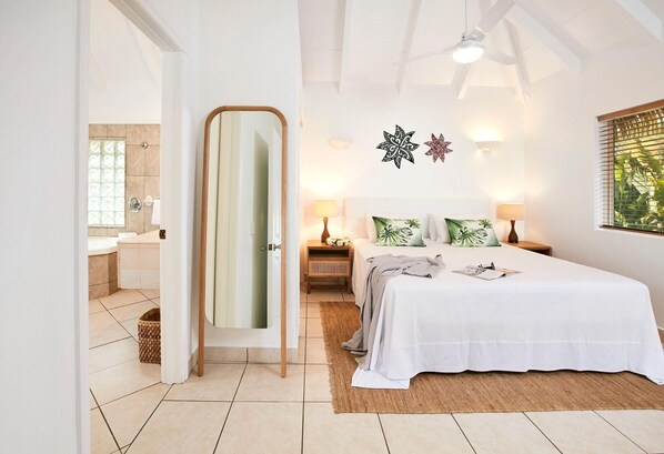 A bedroom with a bed, bedside tables, a mirror, and a window with blinds. - Motu Beachfront Art Villas (Rarotonga)
