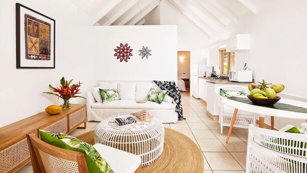 A modern living room with a high ceiling, white walls, and a skylight. There is a white sofa with patterned cushions, a wooden side table with a plant, and a white wicker coffee table. A kitchen area with white cabinets and a dining table with white chairs is visible in the background. - Motu Beachfront Art Villas (Rarotonga)