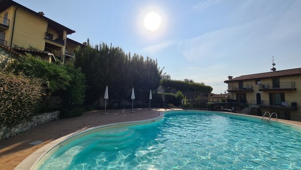 A swimming pool surrounded by buildings, with a clear sky and sun. - Casa il Cielo (Polpenazze del Garda)