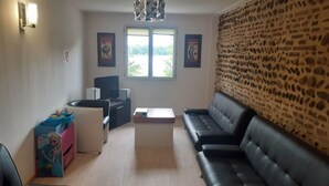 A living room with a black leather sofa, a small table, a TV, and a wall with a stone pattern. There is a window with a view of greenery outside. - Group Lodging and Event Space: La Ferme d'Odette (Poucharramet)