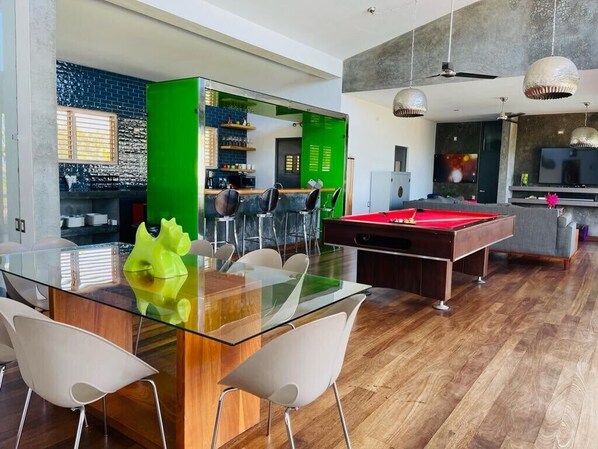 A modern living room with a glass dining table, pool table, and bar area with a green accent wall.