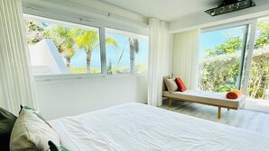 A bedroom with a large bed, a bench with pillows, and a view of trees and sky through large windows. - V12 Casa Summerhill  · Luxury villa w/ 2-level pool + bar and pool table (Sosúa)