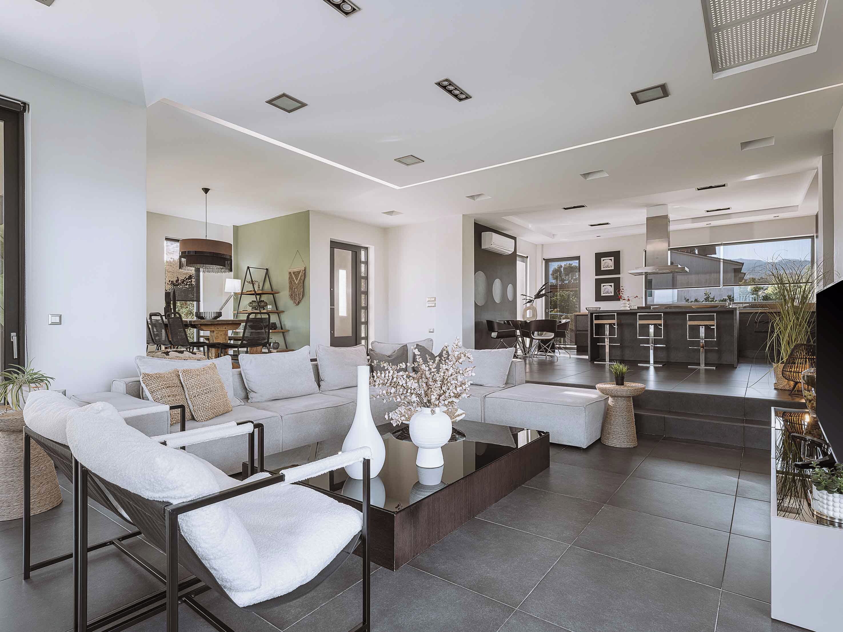 Un salon moderne avec un canapé, une table basse et des fauteuils. On aperçoit un coin cuisine avec une table à manger et des chaises, visibles à travers une porte ouverte.
