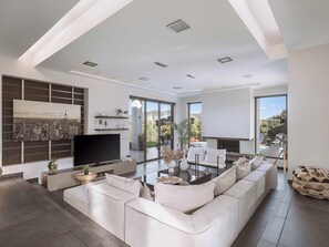 A modern living room with a large sectional sofa, a flat-screen TV, and a cityscape painting on the wall. - Villa Vega Heated* pool, next to nature! (Chania)