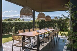 An outdoor dining area with a wooden table and wicker chairs, surrounded by a hedge and a view of mountains. - Villa Vega Heated* pool, next to nature! (Chania)