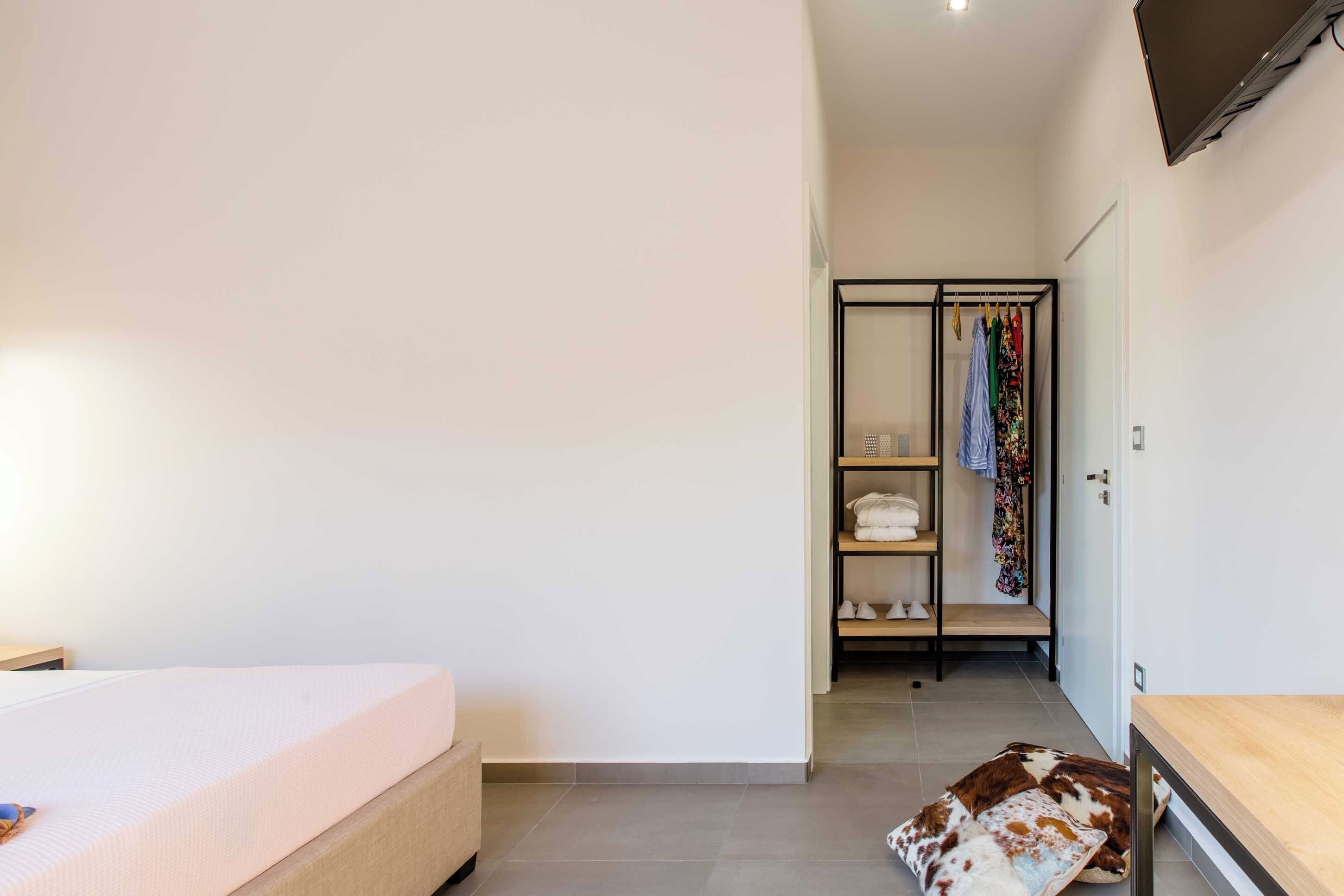 A bedroom with a bed, a wooden nightstand, a closet with hanging clothes, and a small table.