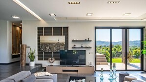 Ein modernes Wohnzimmer mit einem großen Flachbildfernseher, bequemen Sitzgelegenheiten, einem Couchtisch und Blick auf eine gebirgige Landschaft durch große Schiebetüren.